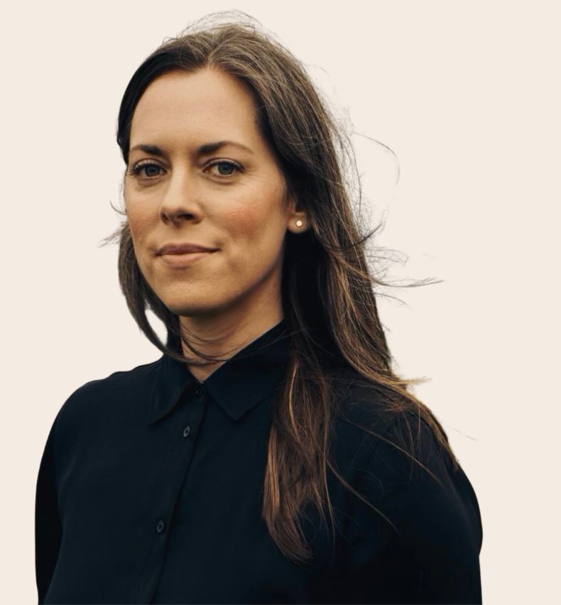 A woman with shoulder-length brown hair wearing a black collared shirt, standing outdoors with wind gently blowing her hair, against a plain, light background.
