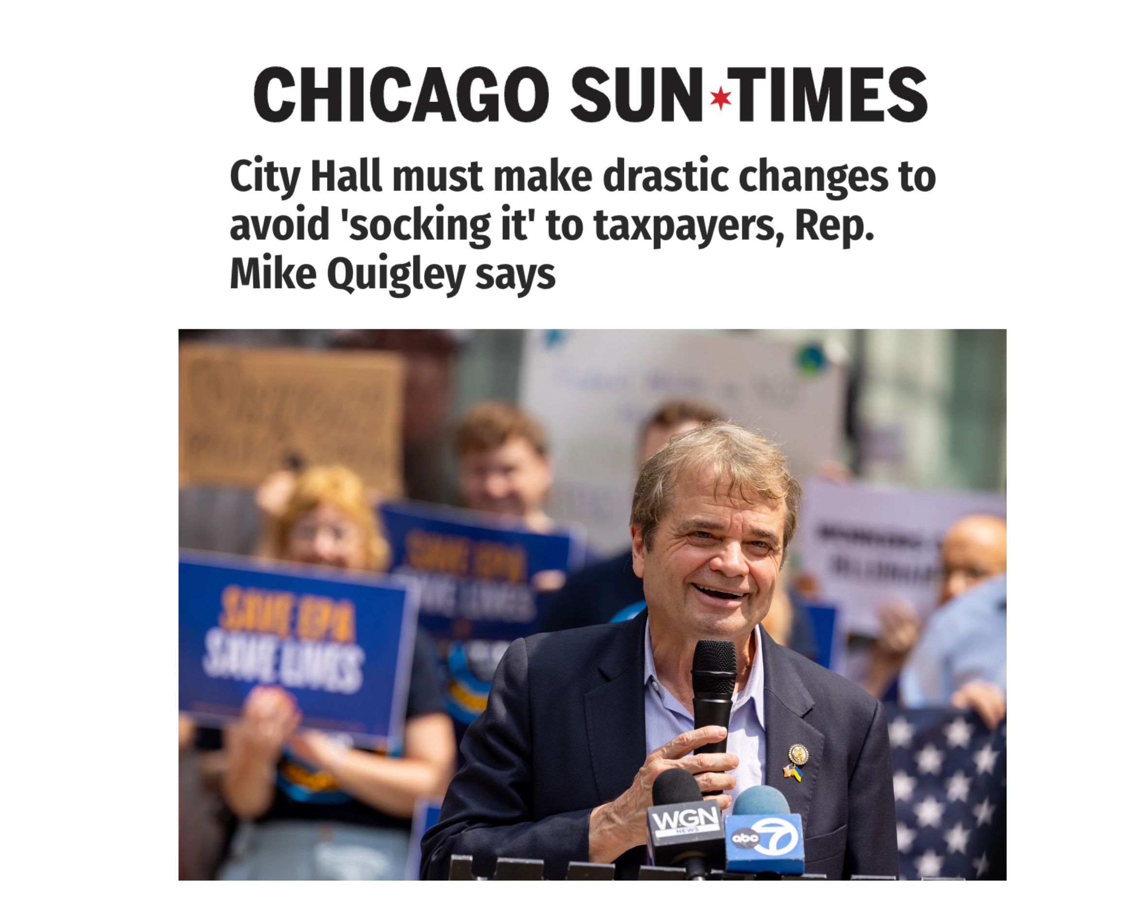 Man speaking into a microphone at a press conference with a crowd holding signs in the background.