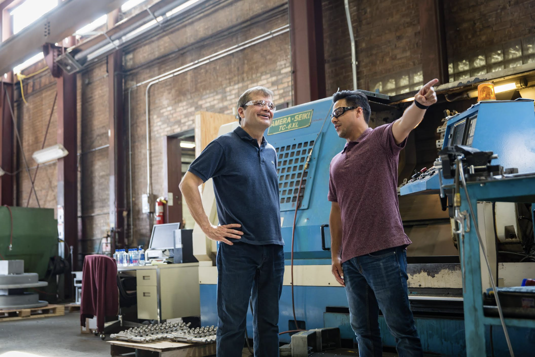 Two men in safety glasses standing in front of a large blue CNC machine in a workshop, with one man pointing and explaining something.