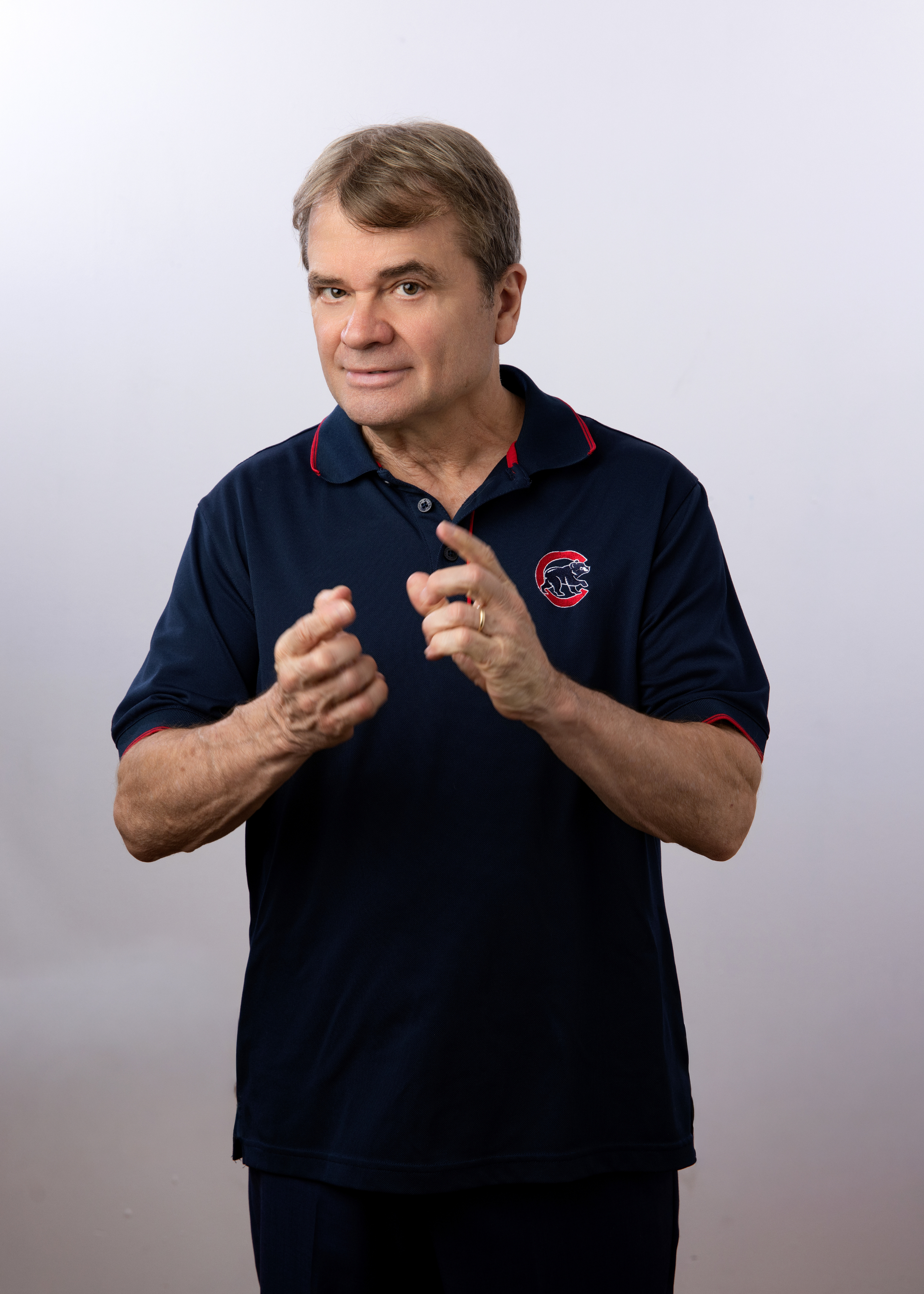 A middle-aged man with short light brown hair pointing with his right index finger towards the camera, wearing a dark blue polo shirt with red accents and a logo of a fierce animal on the left chest, standing against a plain light grey background.