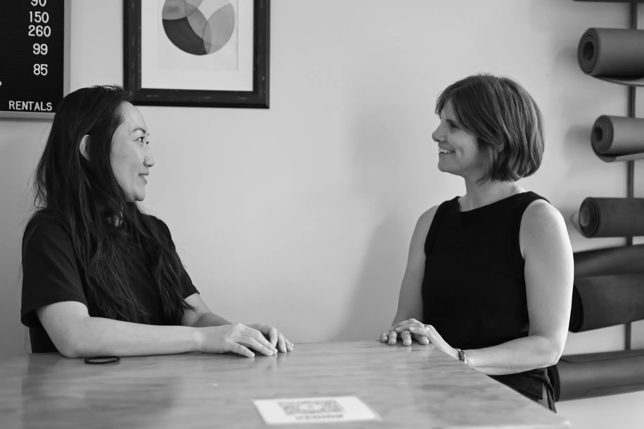 Two women engaged in conversation at a table in a room with artwork and rolled mats on the wall.