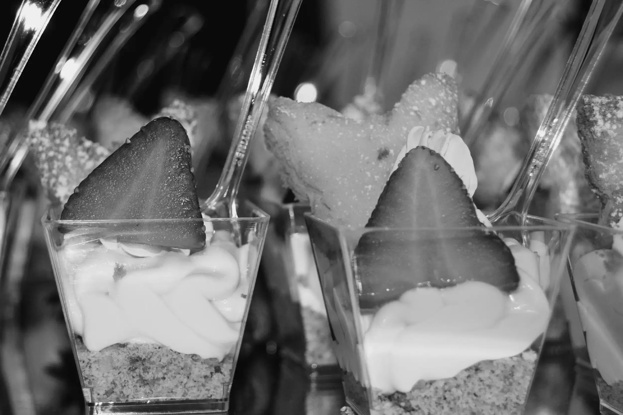 Close-up of several dessert cups filled with whipped cream, crushed cookies, and topped with slices of strawberries, with plastic spoons inserted.