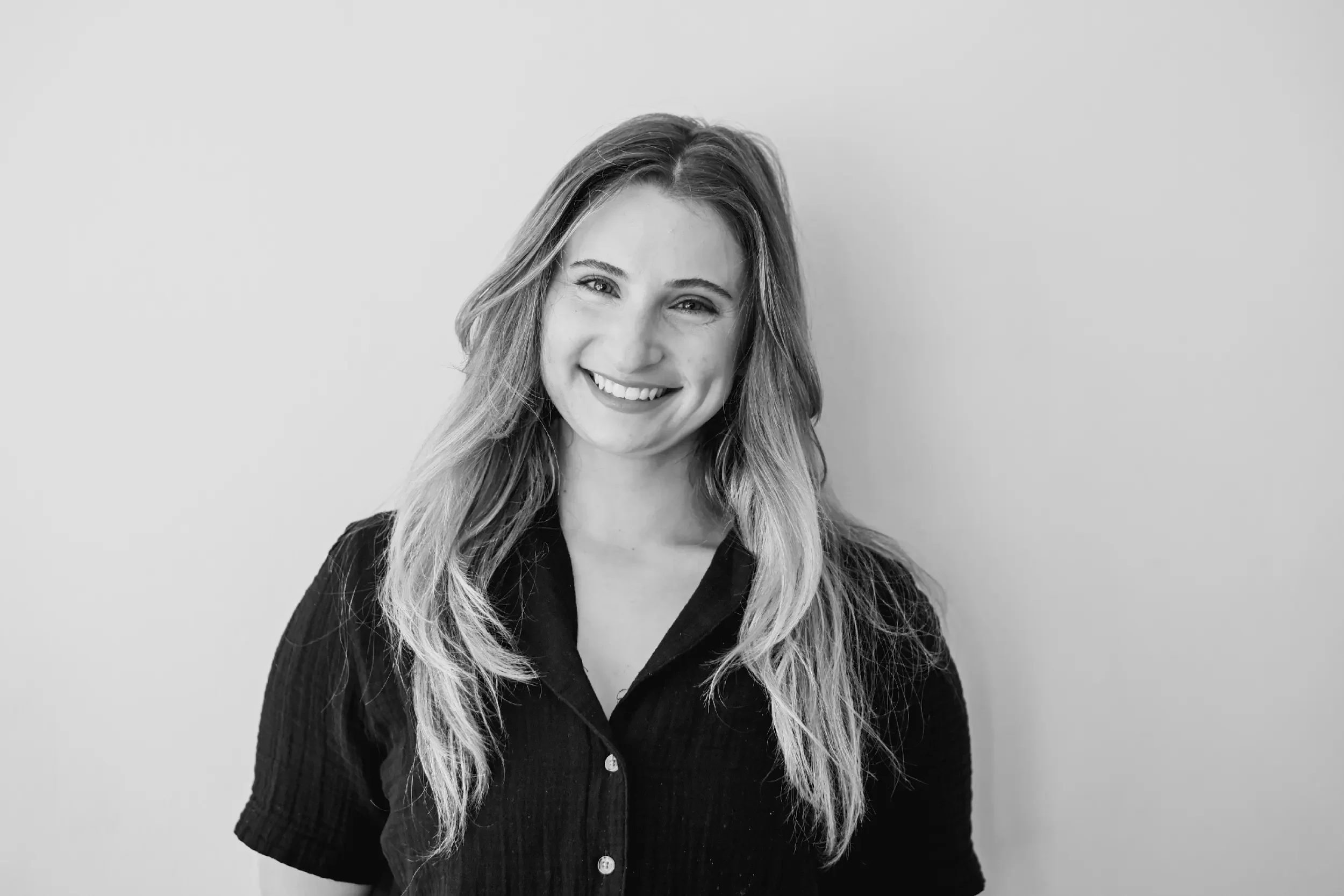 Black-and-white portrait of a smiling woman with long, wavy hair, wearing a dark, short-sleeved, button-up top, standing against a plain light-colored wall.