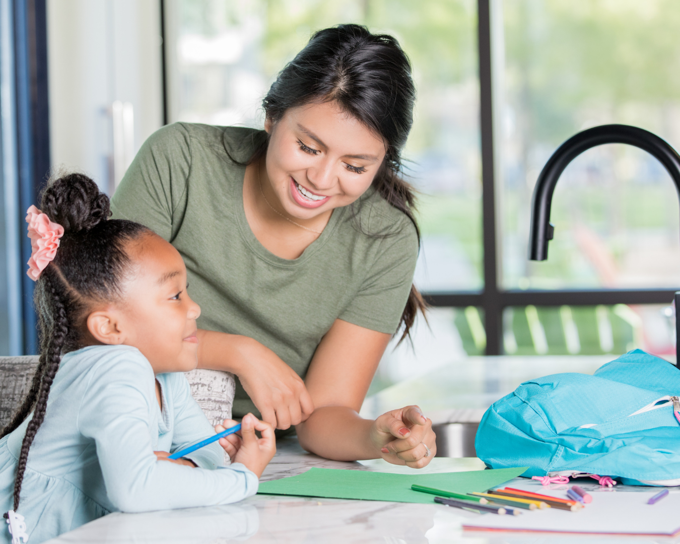 A young woman with dark hair smiling and leaning over a table while helping a young girl with braids hold a blue pen. They are in a bright room with a window and a blue backpack on the table.