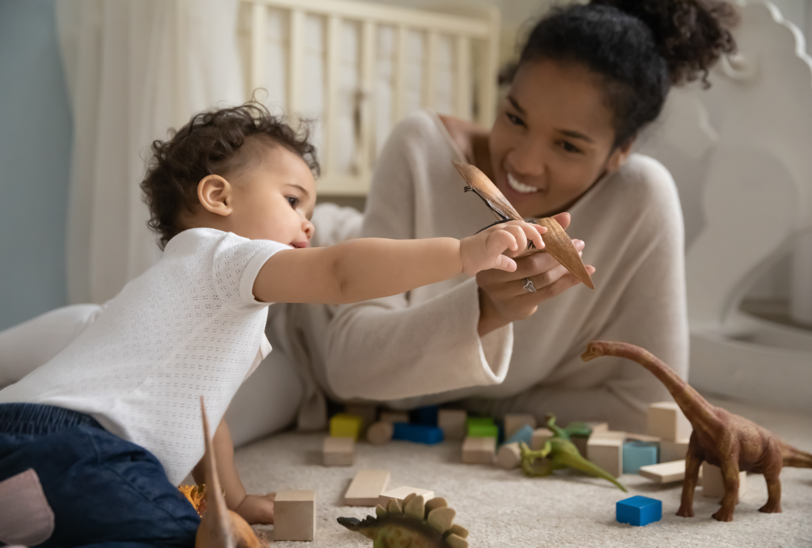 A woman and a young boy playing with toy dinosaurs and blocks on a carpeted floor, sharing a moment of joy and discovery.
