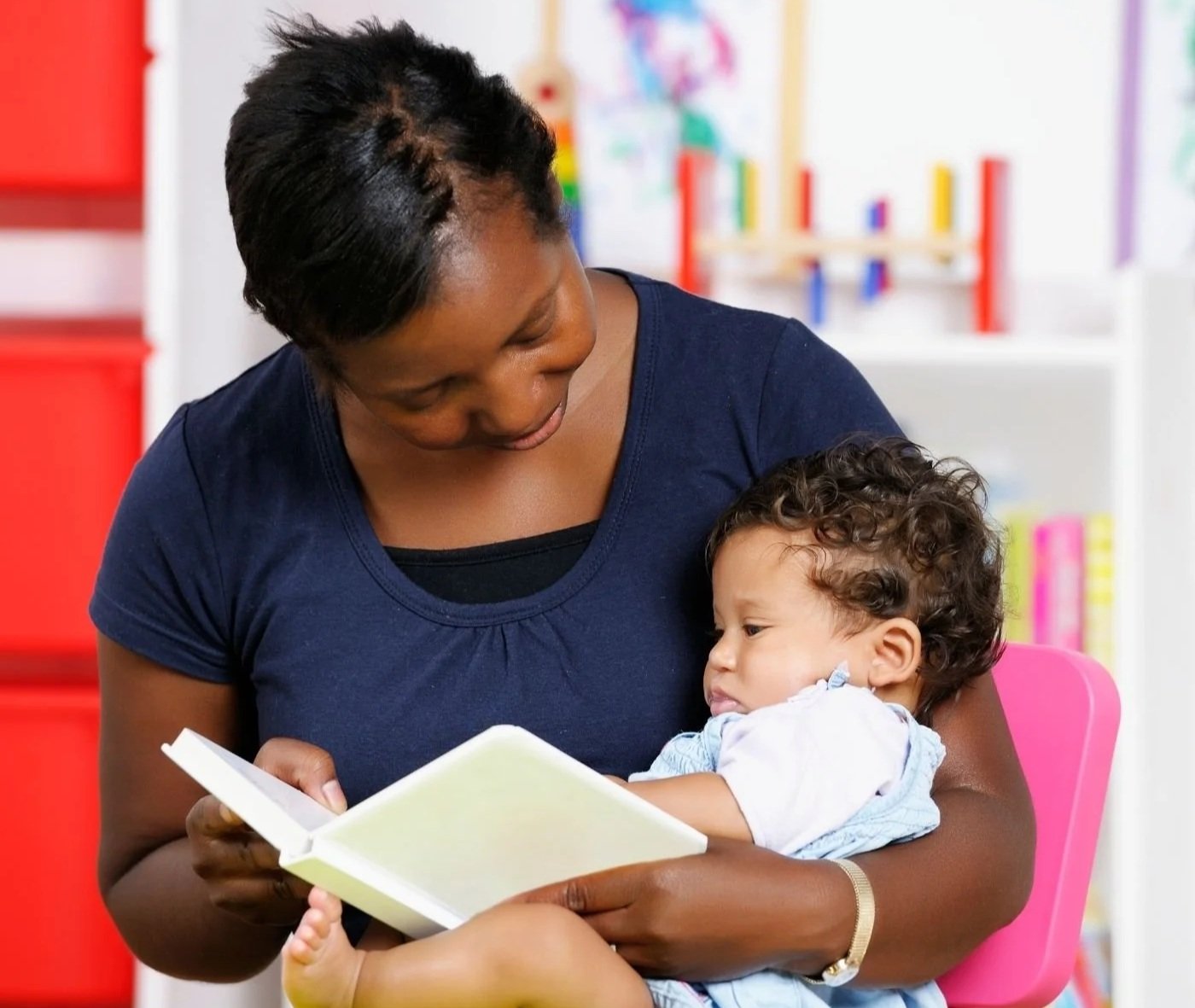 A woman reading a book to a young child sitting on her lap, in a colorful room with bookshelves and educational toys in the background.