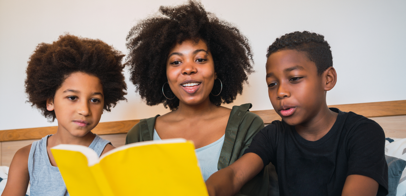 A woman and two children reading a yellow book together on a bed in a bedroom.
