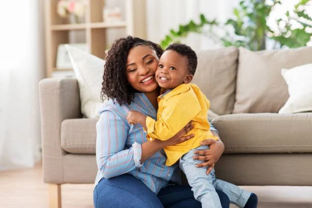 Woman and young boy hugging and smiling in a living room with a sofa and green plant in the background.