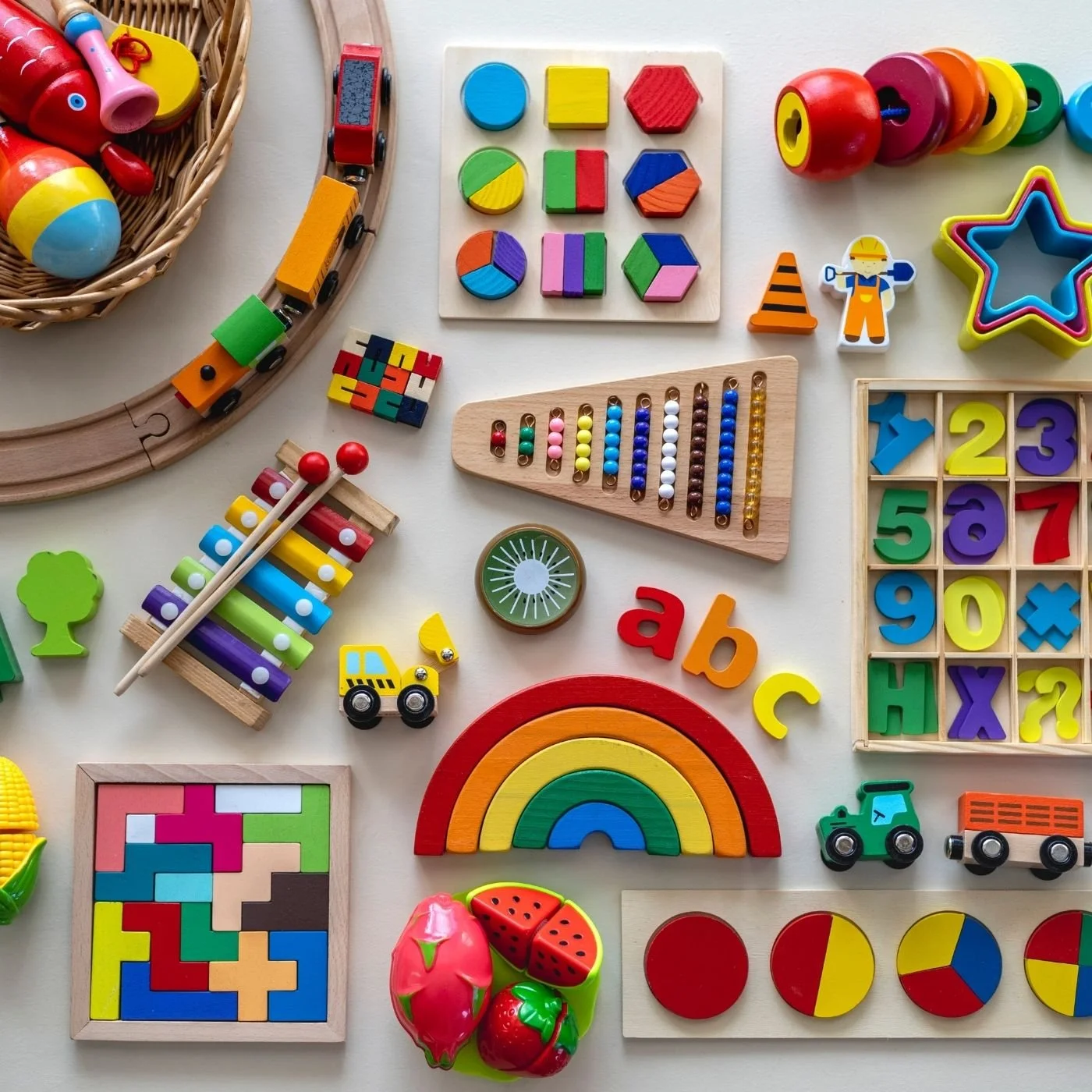 Colorful children's toys and educational game pieces scattered on a white surface, including a rainbow stacker, wooden train, abacus, puzzle, alphabet and number blocks, and toy fruits.
