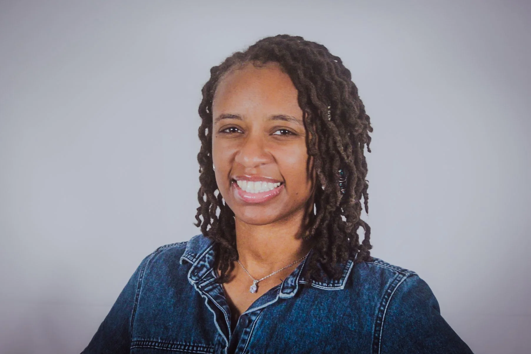 A woman with shoulder-length dreadlocks smiling and wearing a denim jacket and a silver necklace against a plain light gray background.
