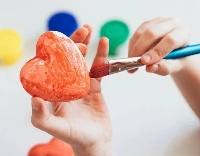Person painting a red, heart-shaped object with a paintbrush in front of colorful blurred background.