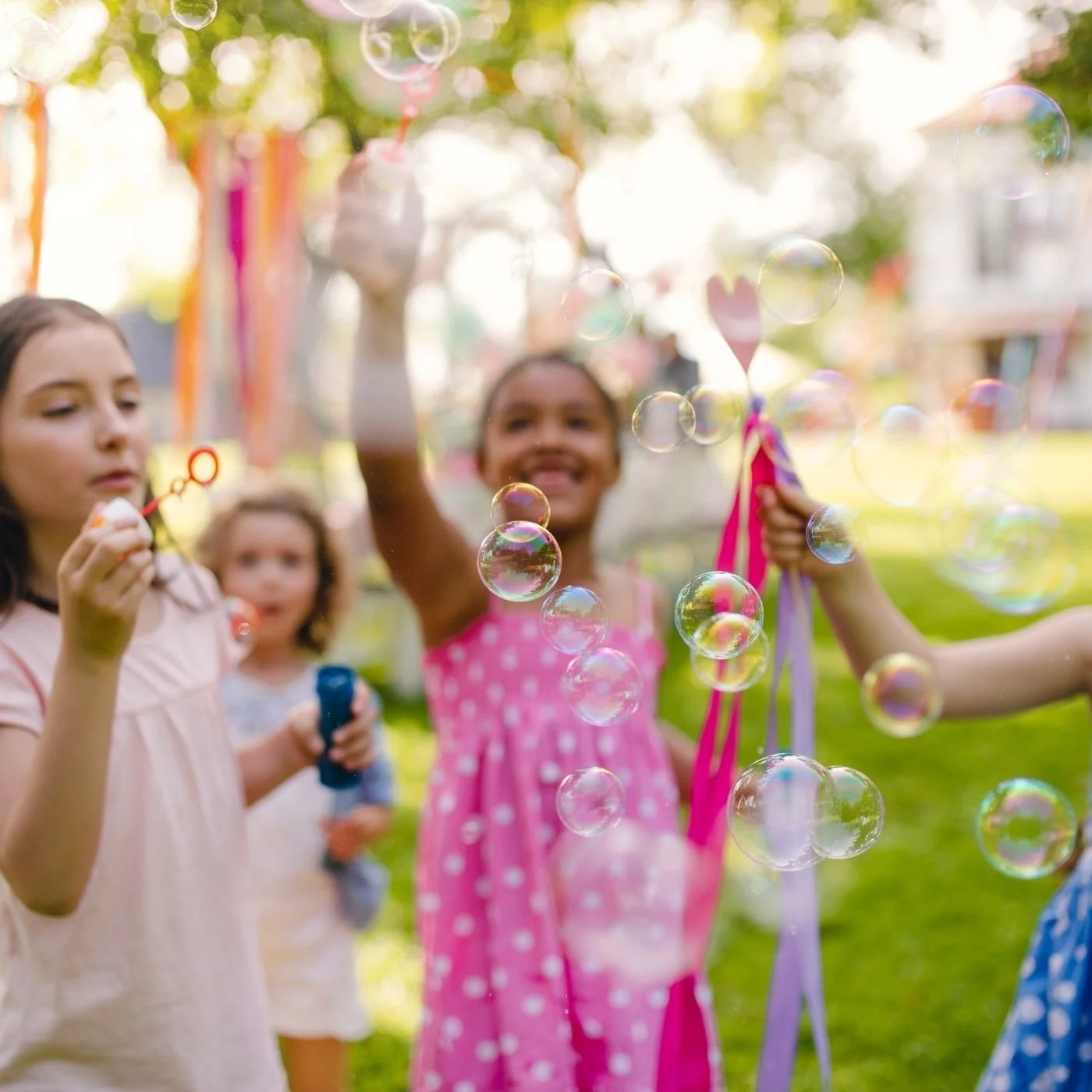 Children playing with bubbles outdoors on a sunny day, with some wearing dresses and holding bubble wands.