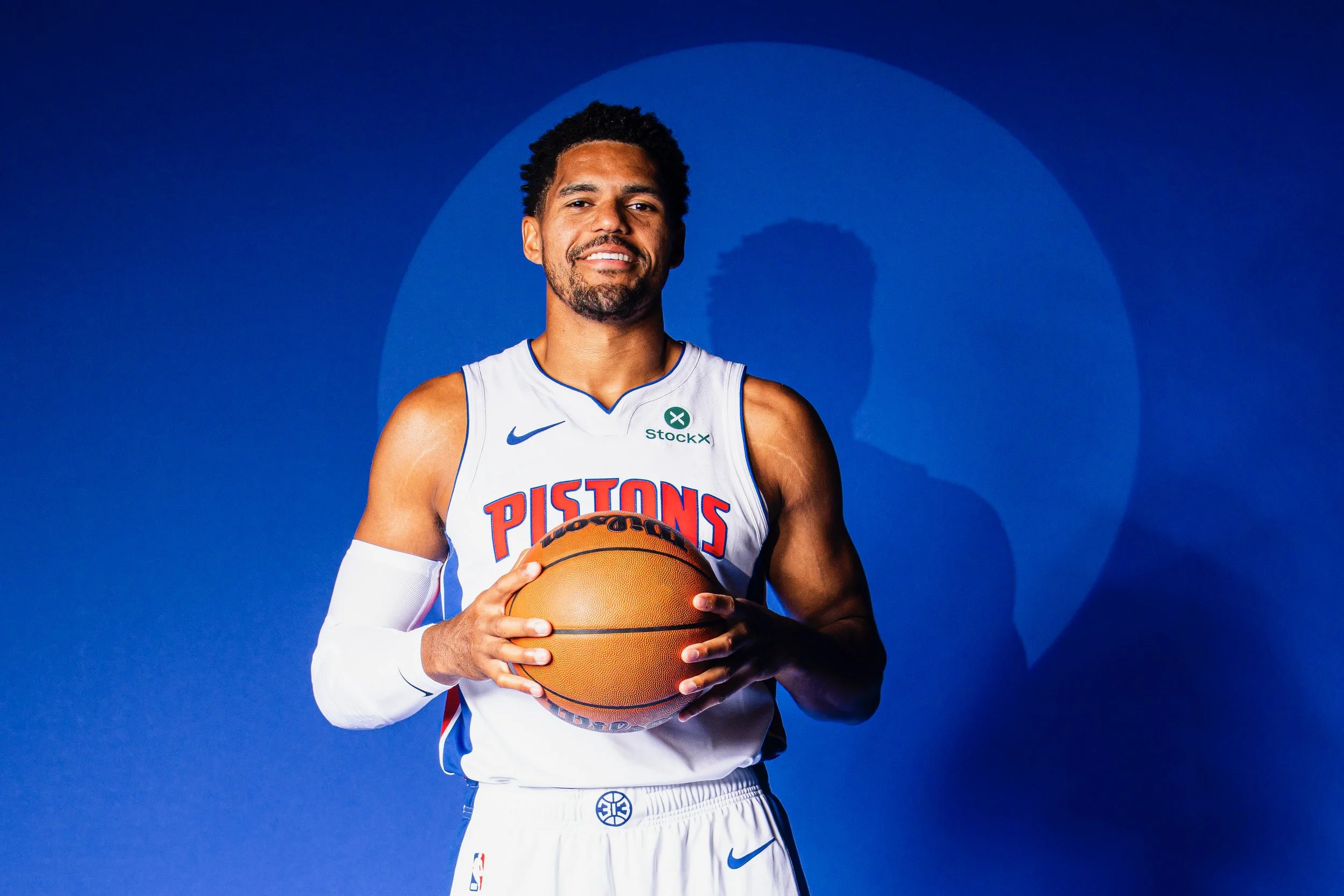 Professional basketball player, Tobias Harris, in a Detroit Pistons uniform holding a basketball against a blue background.