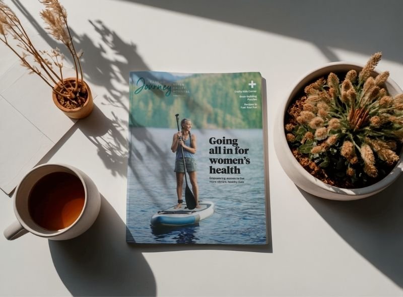 A magazine titled 'Journey' with the headline 'Going all in for women's health,' placed on a white surface alongside a cup of tea, a small plant with pampas grass, and a potted cactus.