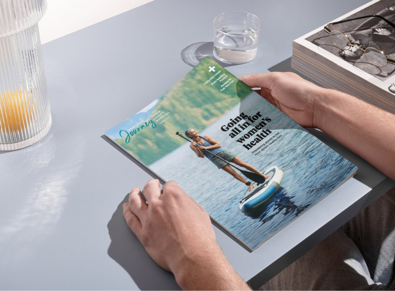 A person holds a magazine titled "Healthy Journey" with a cover image of a woman paddleboarding on a lake, demonstrating theme of women's health.