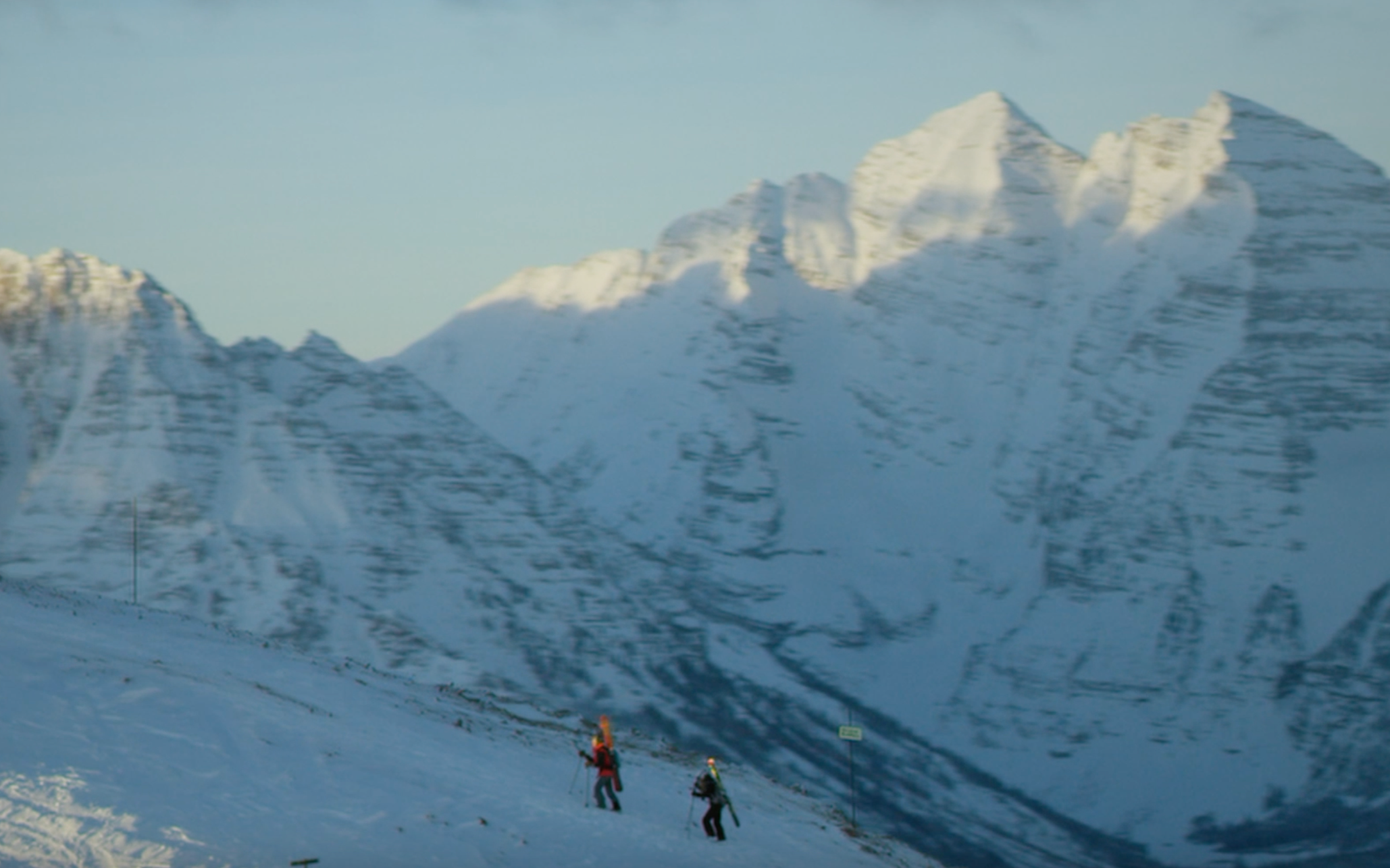 Two skiers in brightly colored clothing skiing in a snow-covered mountain landscape with towering snowy peaks in the background.
