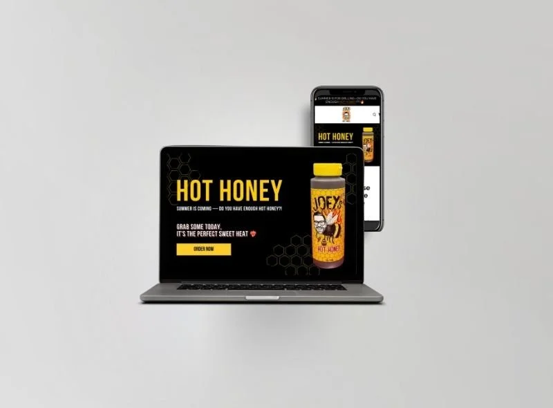 Laptop and smartphone displaying an advertisement for hot honey with a jar and a label featuring a cartoon character, promoting its summer availability and sweet heat.