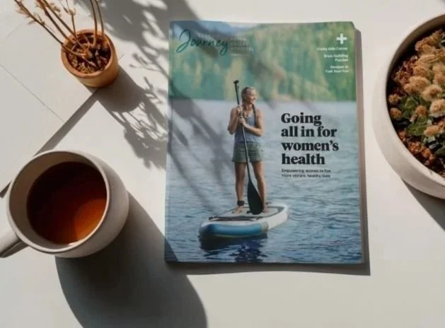 Magazine titled "Healthy Journey" on a table with a cover image of a woman paddleboarding on a lake, along with a white cup of tea or coffee, a small plant in a brown pot, and a larger plant in a white pot.