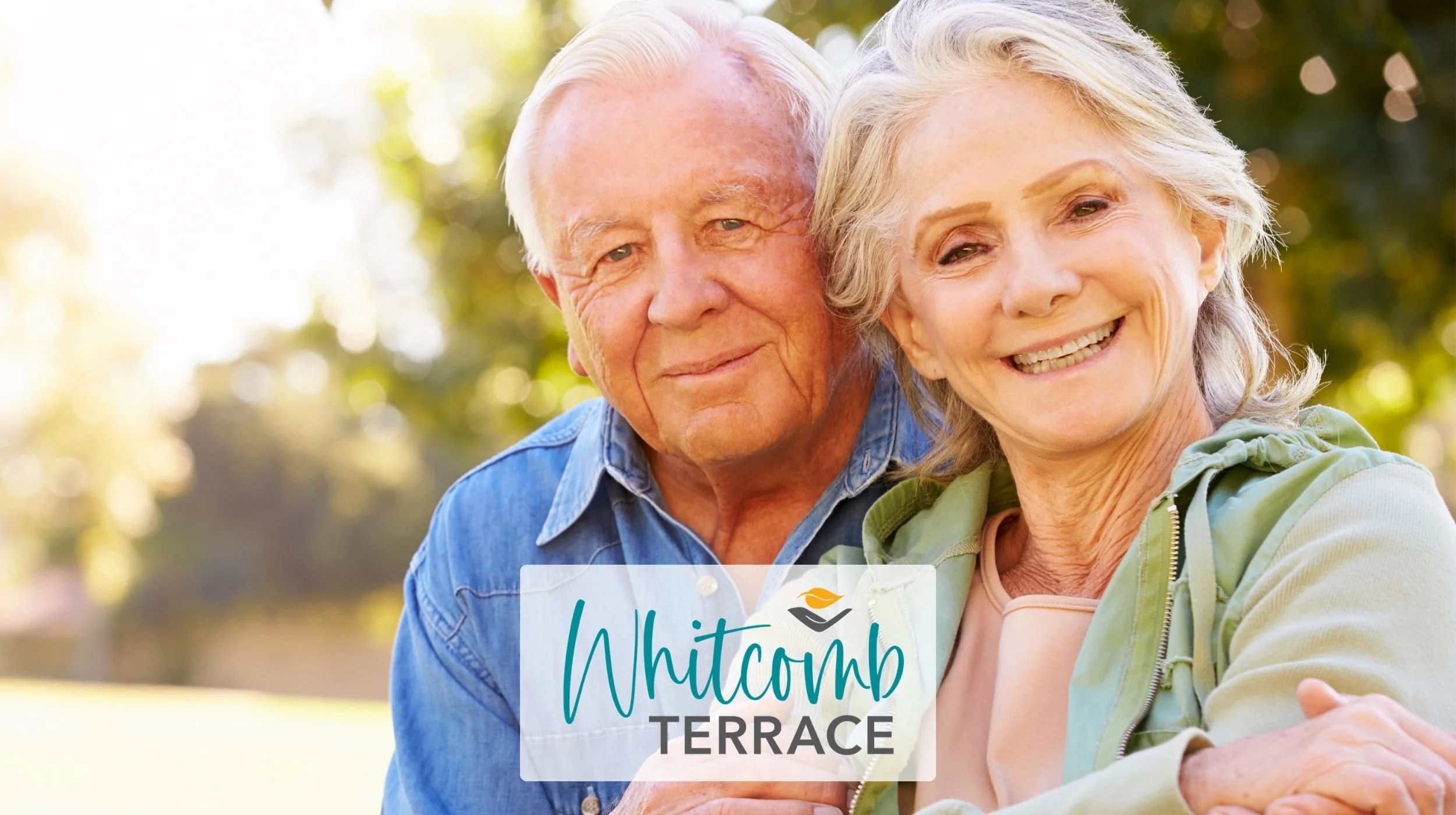 A photo of a senior couple huggin as they smile for the camera. The Whitcomb Terrace logo is superimposed at the bottom center of the photo.