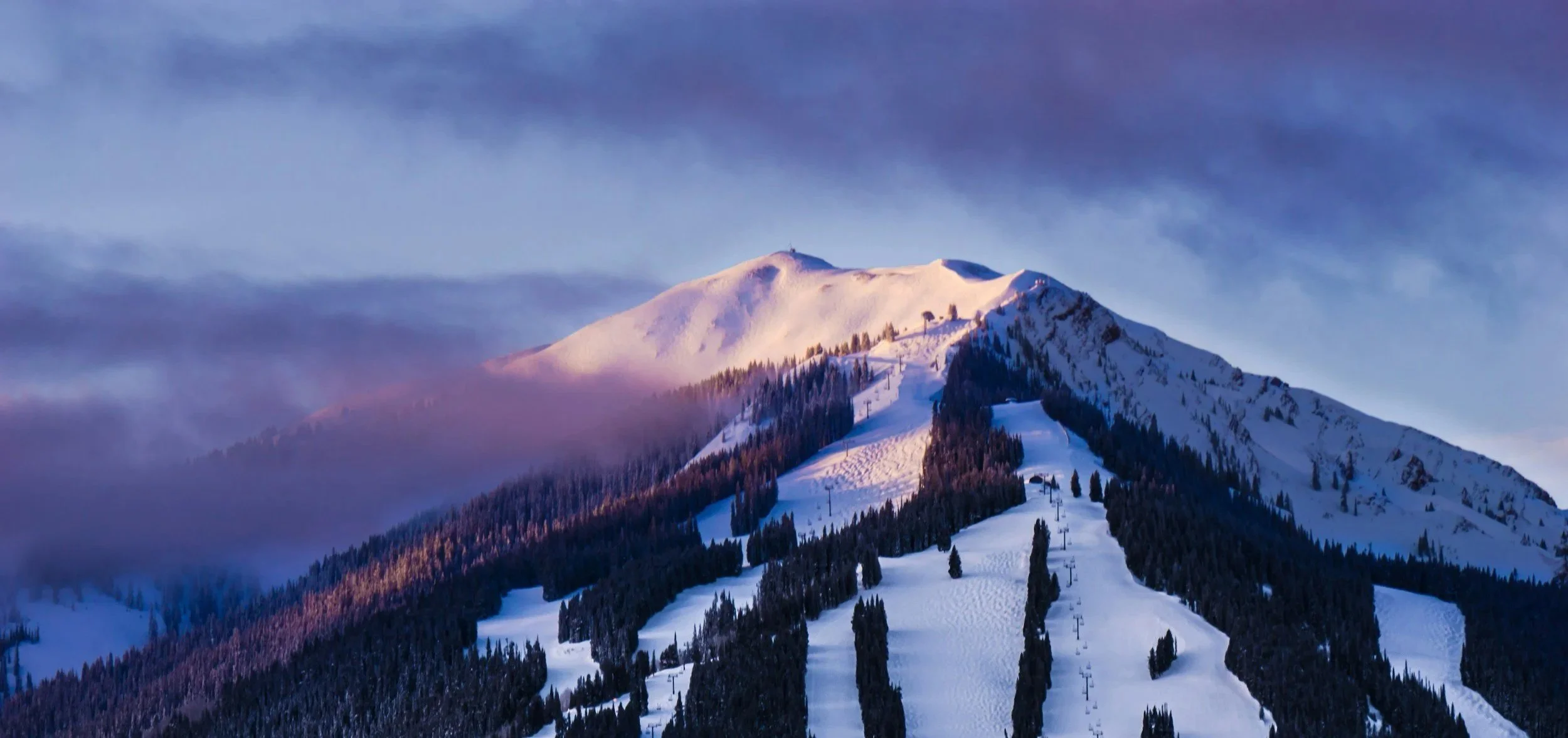 Snow-covered mountain with ski slopes, ski lifts, and tall pine trees against a partly cloudy sky at sunset.