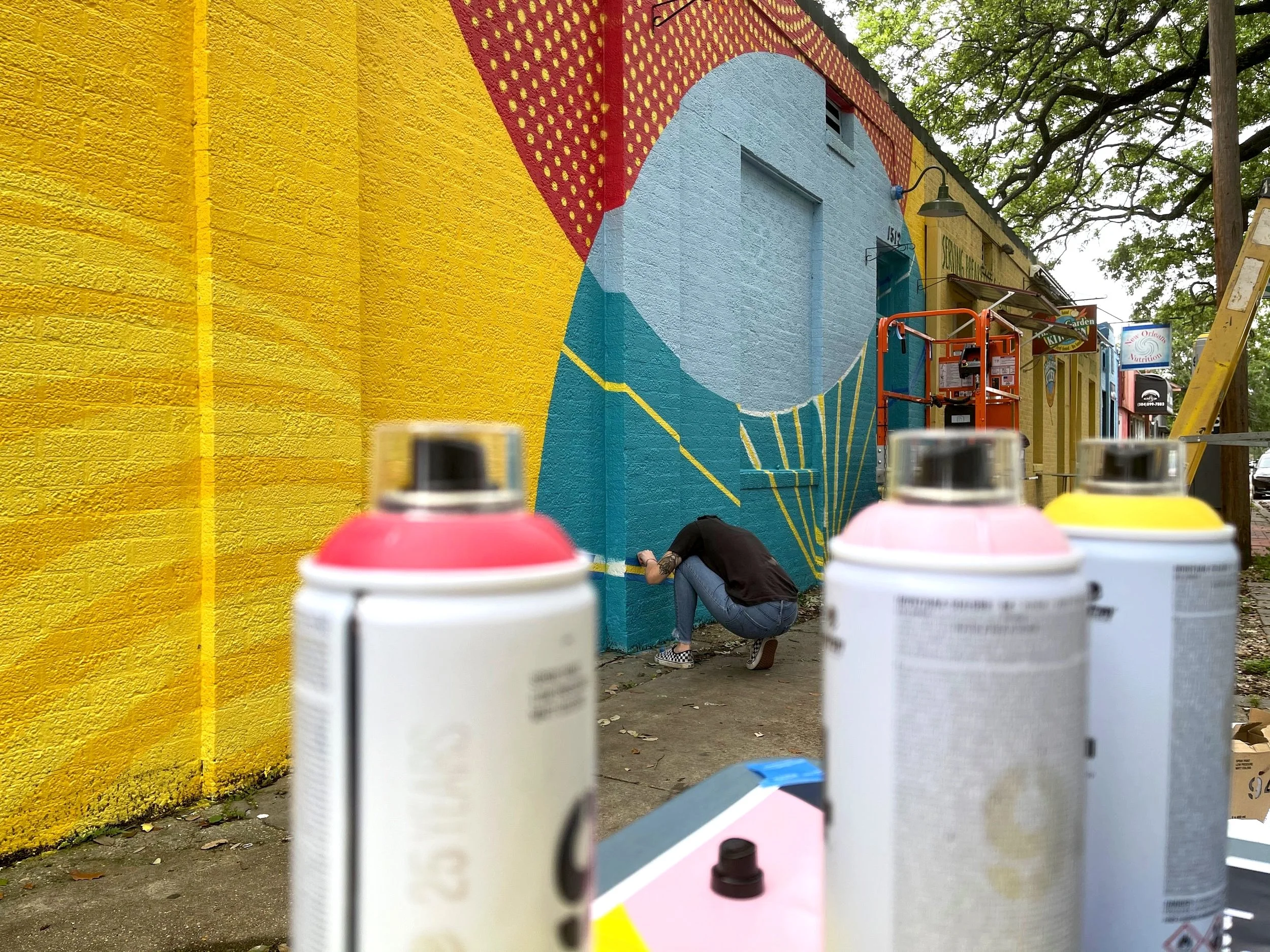 A person crouched down spray painting a mural on a building with a yellow wall, while in the foreground there are four cans of spray paint in pink, yellow, and white.