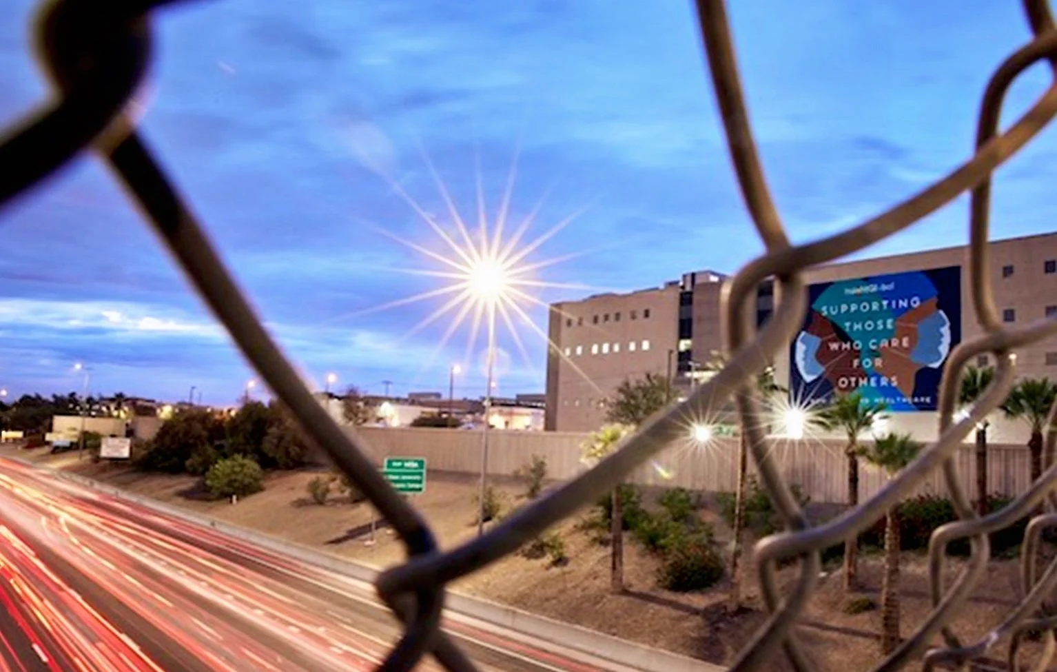 City street view taken through a chain-link fence at dusk, showing light trails from moving vehicles, a billboard with a message about supporting those who care for others, streetlights, and a large building in the background.