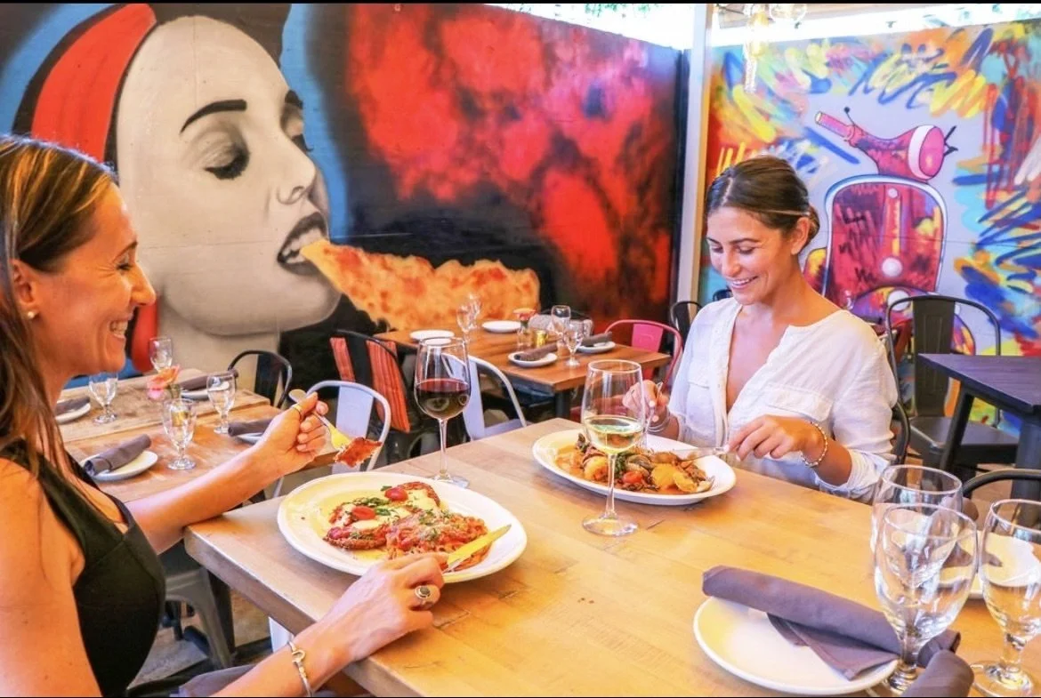 Two women enjoying pizza and wine at a restaurant with colorful graffiti murals in the background.