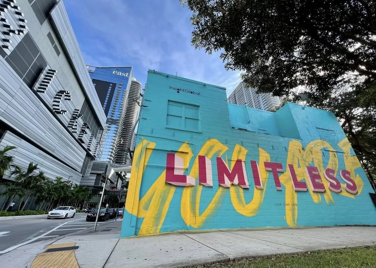 A colorful graffiti mural on a building wall with the words 'LIMITLESS' in large pink letters, with a blue background, yellow design, and the artist's signature 'LOVE, LIKE, JON' at the bottom. Surrounding urban city buildings and a street scene with