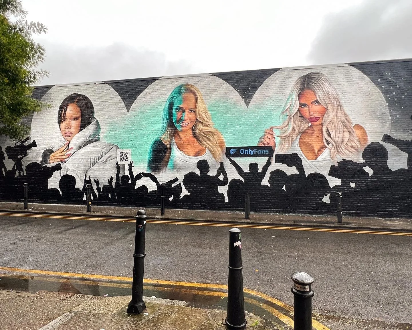 Mural of three women with varying hairstyles and expressions, with the 'OnlyFans' logo and a QR code, set against a cloudy sky.