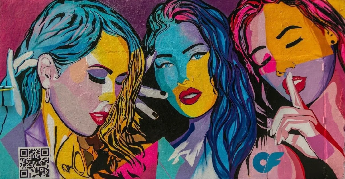 Colorful mural of three women with vibrant hair, the one on the left with yellow hair, the middle with blue hair, and the right with pink hair, all making shushing gestures with their fingers.