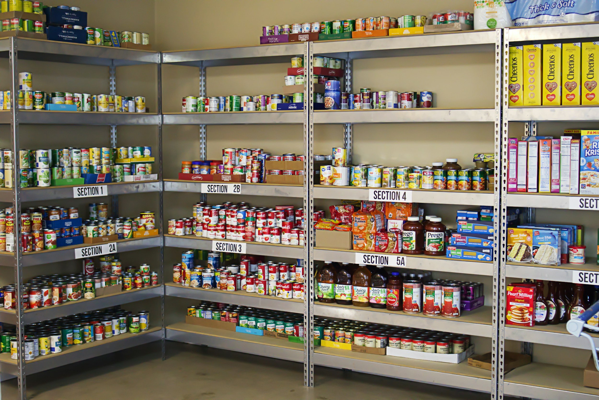 Shelves stocked with canned foods, boxed cereals, and canned juices, labeled with sections 1 through 5A in a food pantry or storage room.
