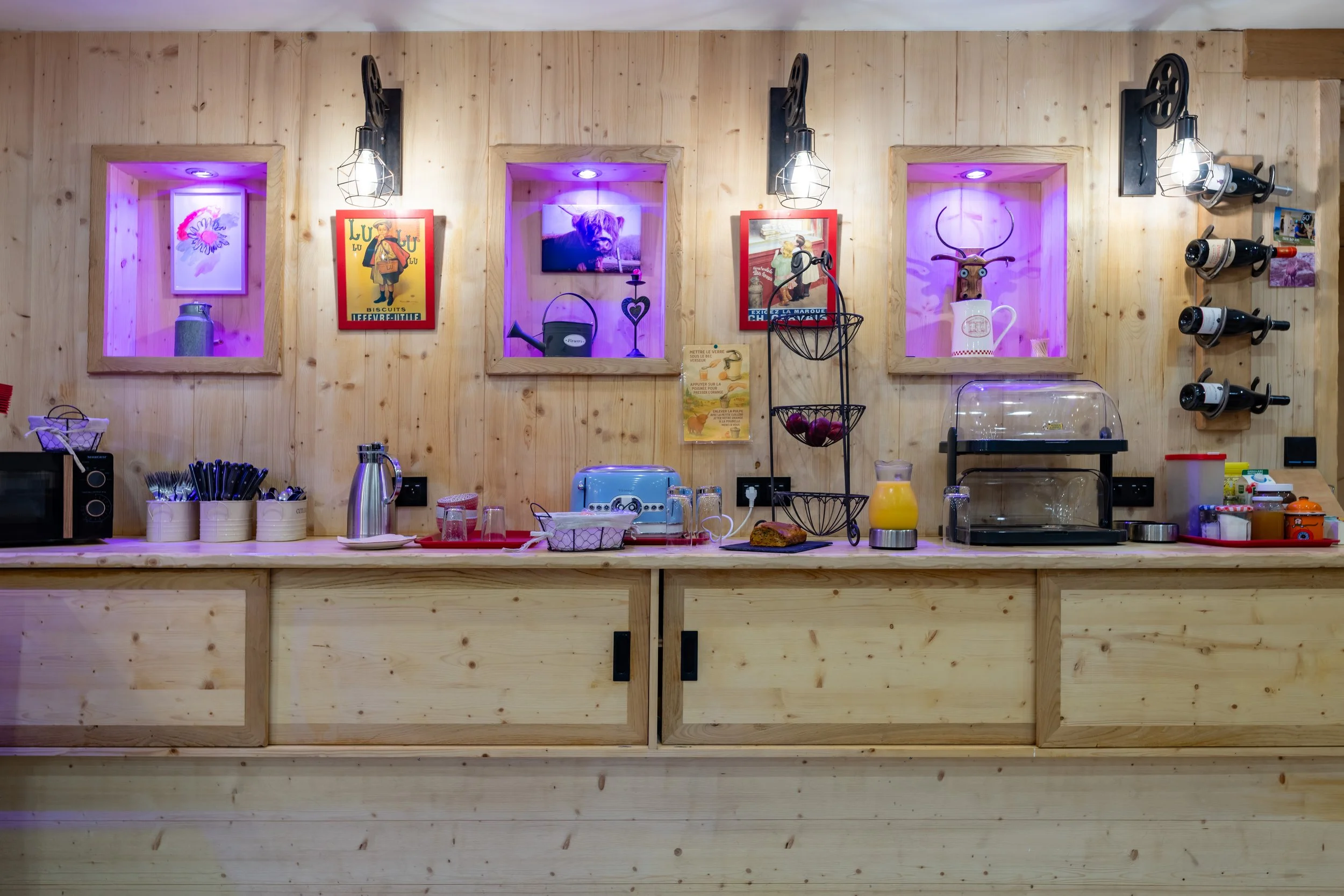 Comptoir de petit déjeuner en bois clair avec diverses machines et objets, mur en bois avec trois niches éclairées en violet contenant des décorations, posters et une télévision, bouteille de jus, vaisselle, toaster, beverage et condiments.