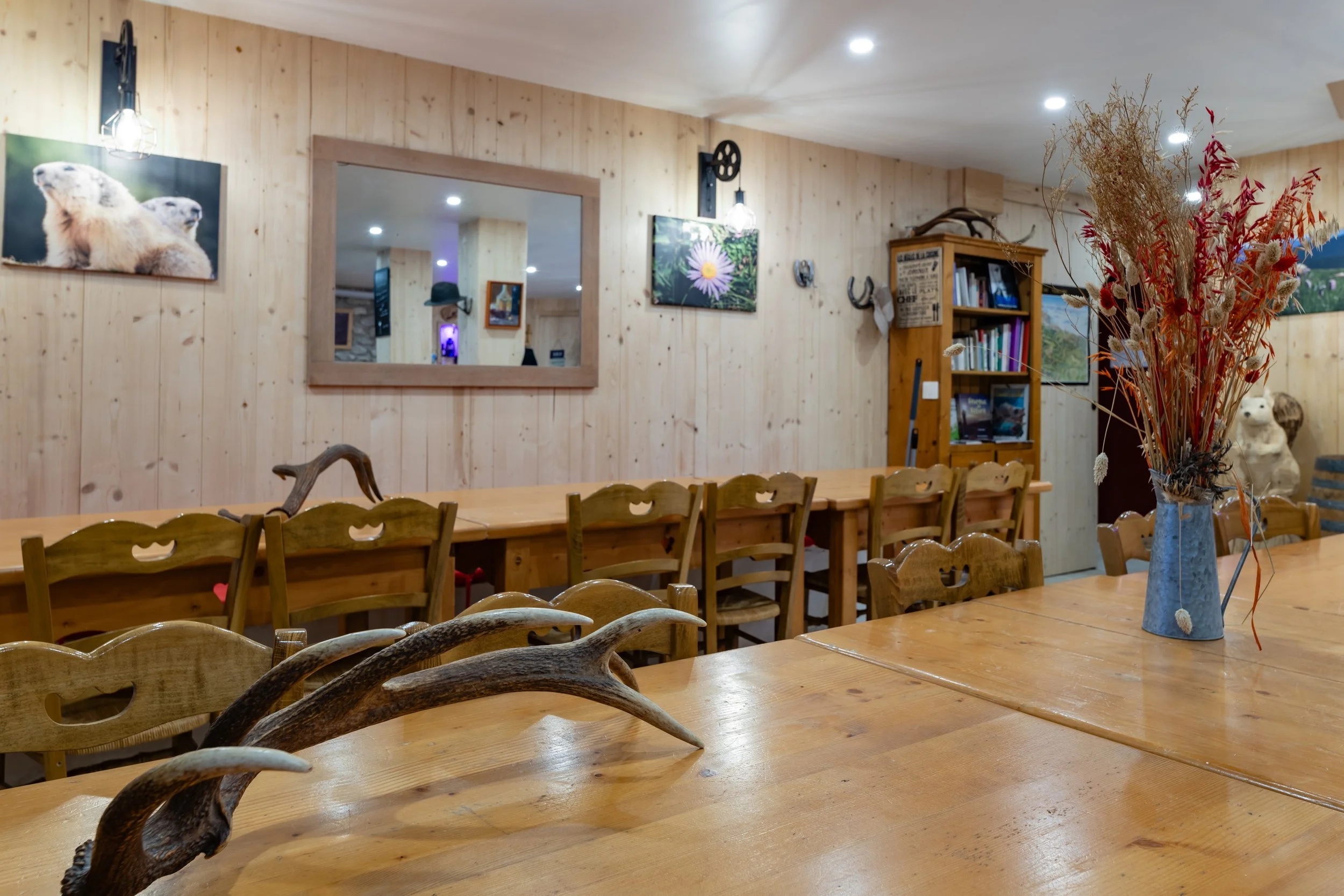 Salle à manger avec des tables en bois, un vase avec des fleurs séchées, un mur en bois clair décoré de photos et d'objets divers, un grand miroir, des livres sur une étagère, et décorations de style rustique.