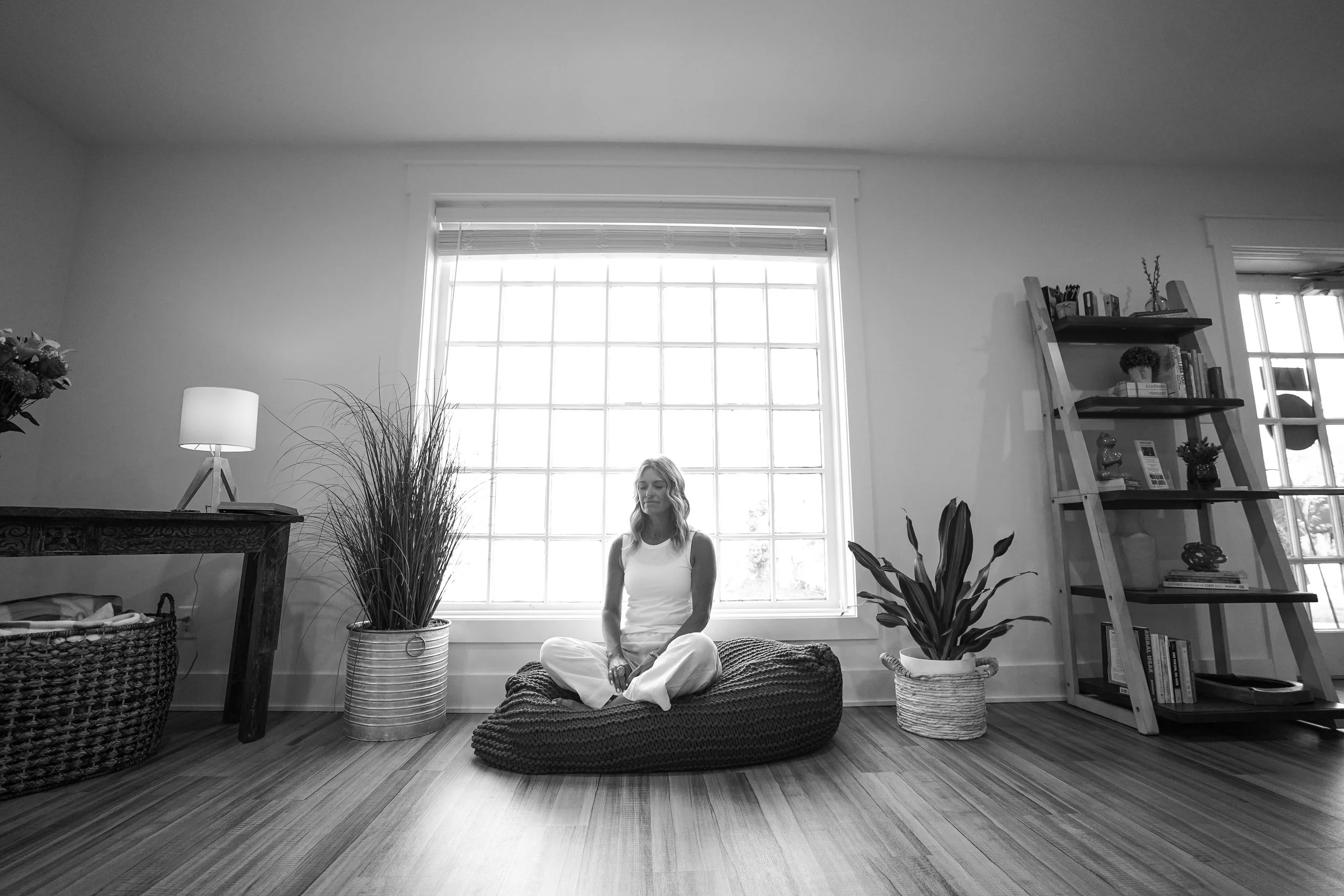 A woman sitting cross-legged on a bean bag in front of a large window in a living room. The room has plants, a bookshelf, and a side table with a lamp.