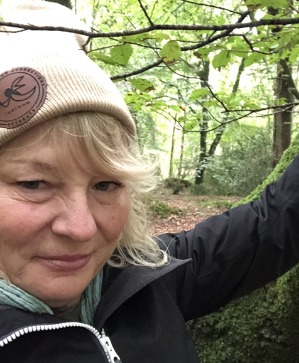 A woman taking a selfie outdoors in a wooded area, wearing a beige beanie and a black jacket.