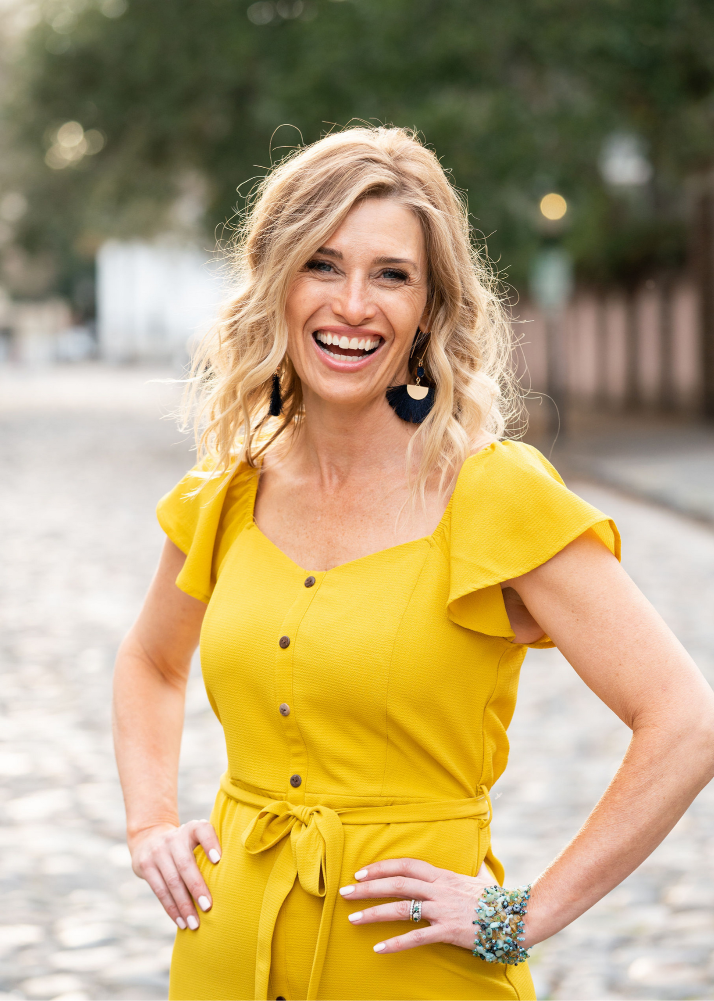 A woman with blonde curly hair, wearing a yellow dress with buttons and a waist tie, standing outdoors on a cobblestone street, smiling and looking at the camera.