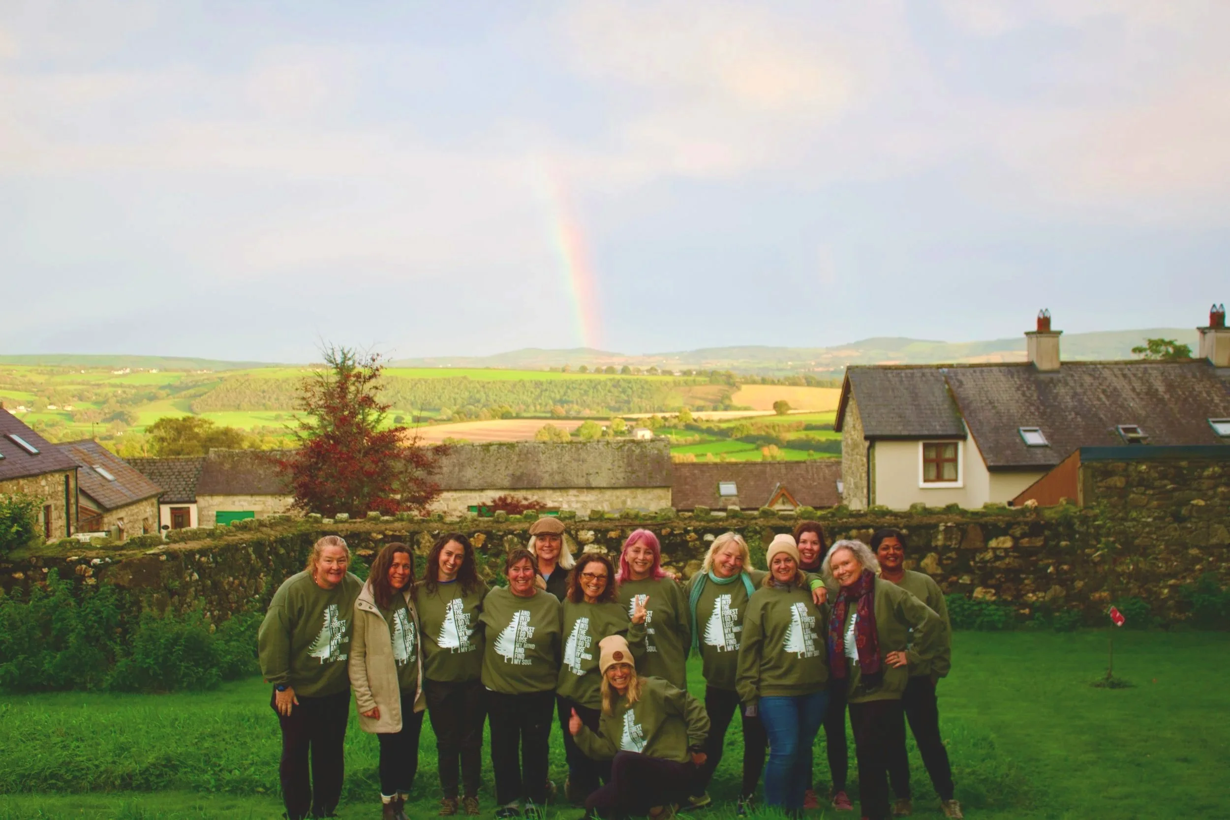 A group of people wearing matching green sweatshirts stands together in a grassy backyard, behind a stone wall, with a rural landscape and a rainbow in the sky in the background.