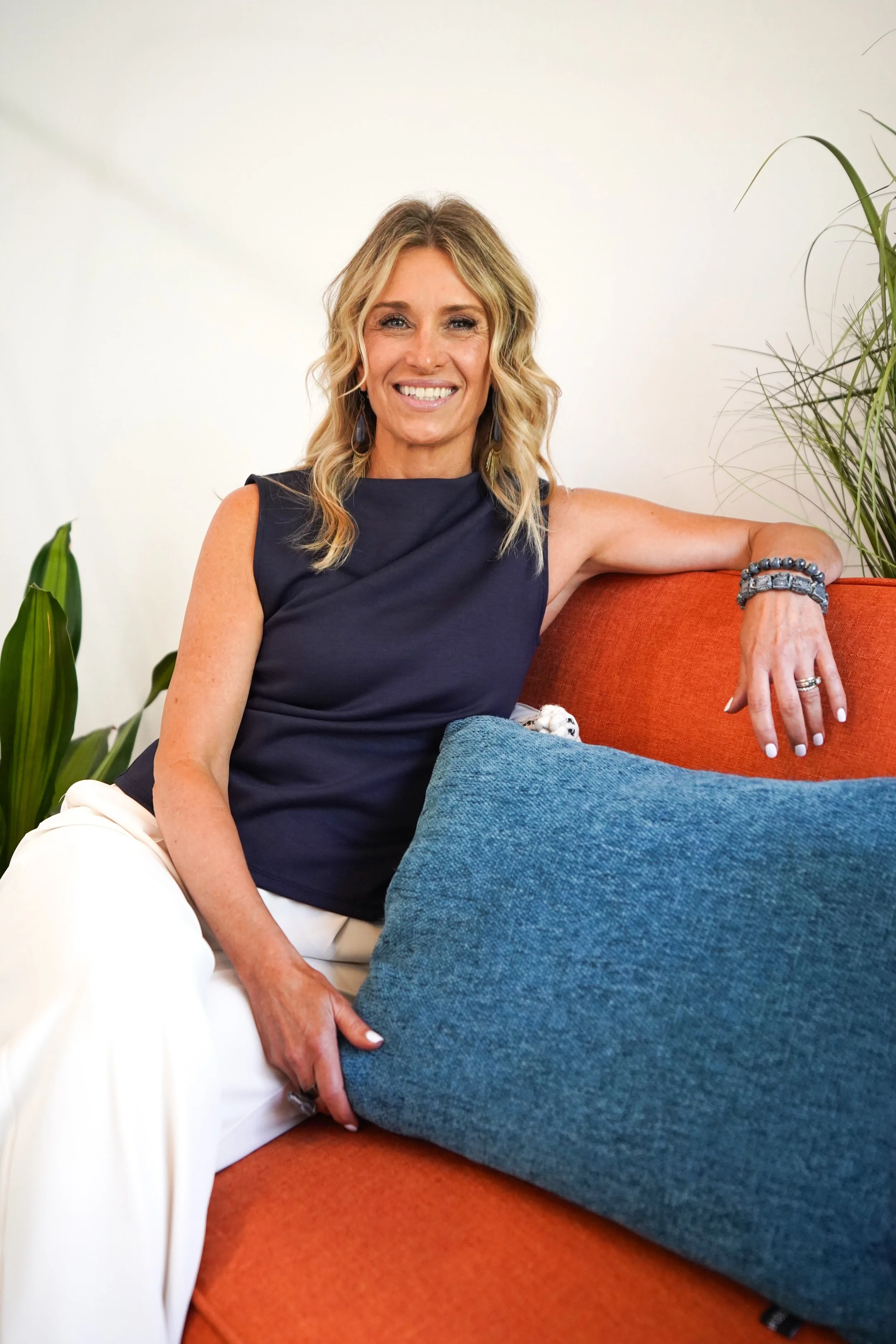 A smiling woman with blonde wavy hair sitting on an orange couch in a room with plants, wearing a sleeveless navy blue top and white pants, with her left arm resting on a large blue pillow.