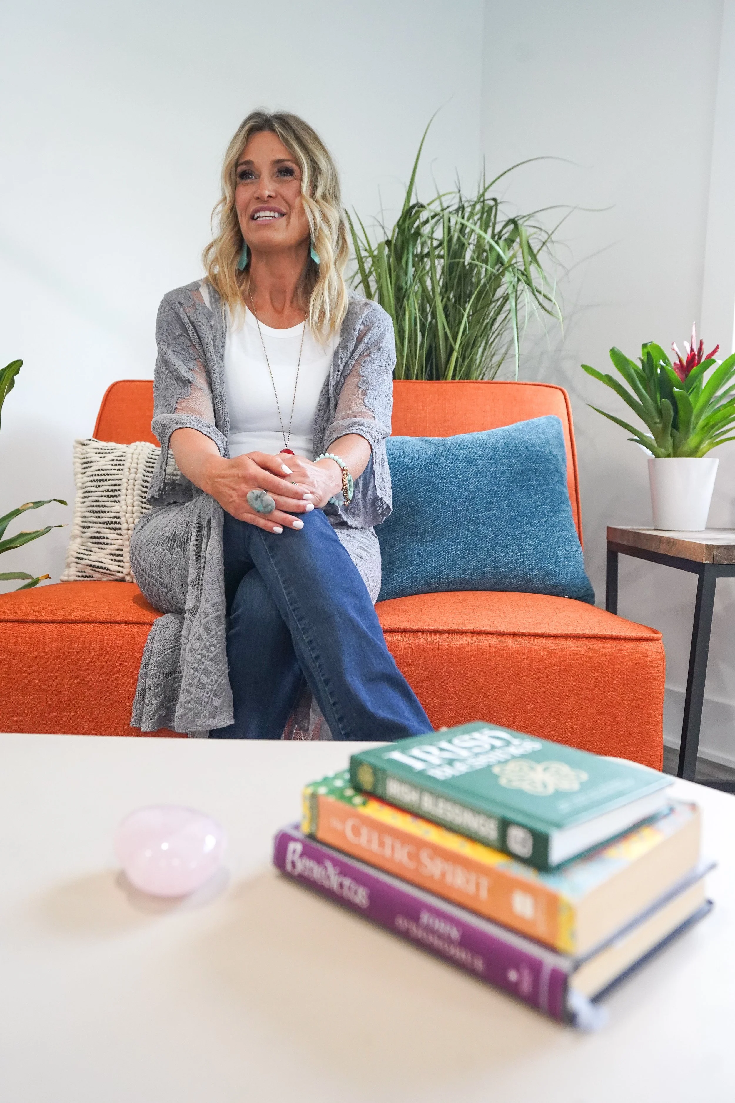 A woman with blonde hair sitting on an orange couch with a blue pillow behind her, in a room decorated with plants. There is a stack of books and a pink decorative object on a white table in the foreground.