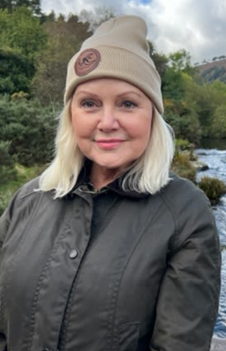 A woman with blonde hair, wearing a beige beanie with a logo and a dark jacket, stands outdoors near a river and greenery.