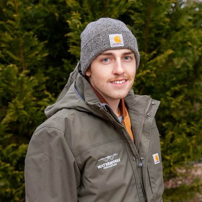 A young man outdoors wearing a gray beanie, olive green Carhartt jacket with a waterworks irrigation logo, and smiling at the camera, surrounded by green trees.