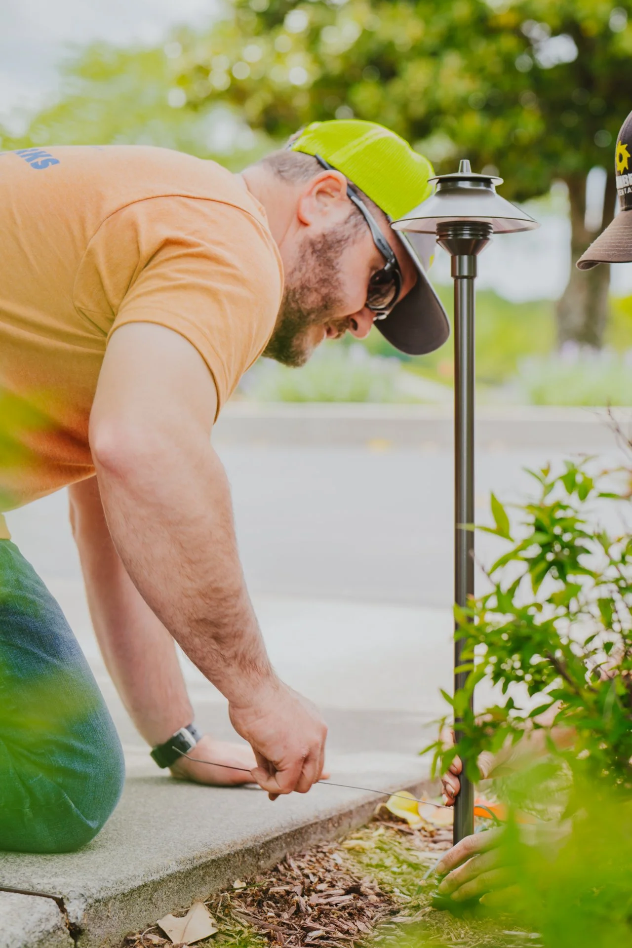A man kneeling on the ground, planting a small tree or shrub outdoors with tools and equipment, wearing sunglasses, a lime green cap, and an orange t-shirt.
