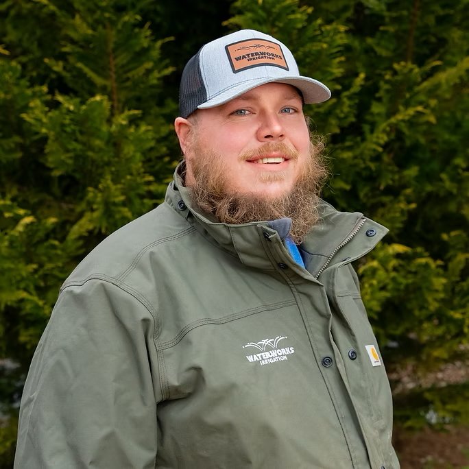 Man with beard wearing a green jacket and cap, standing outdoors with green trees in the background.
