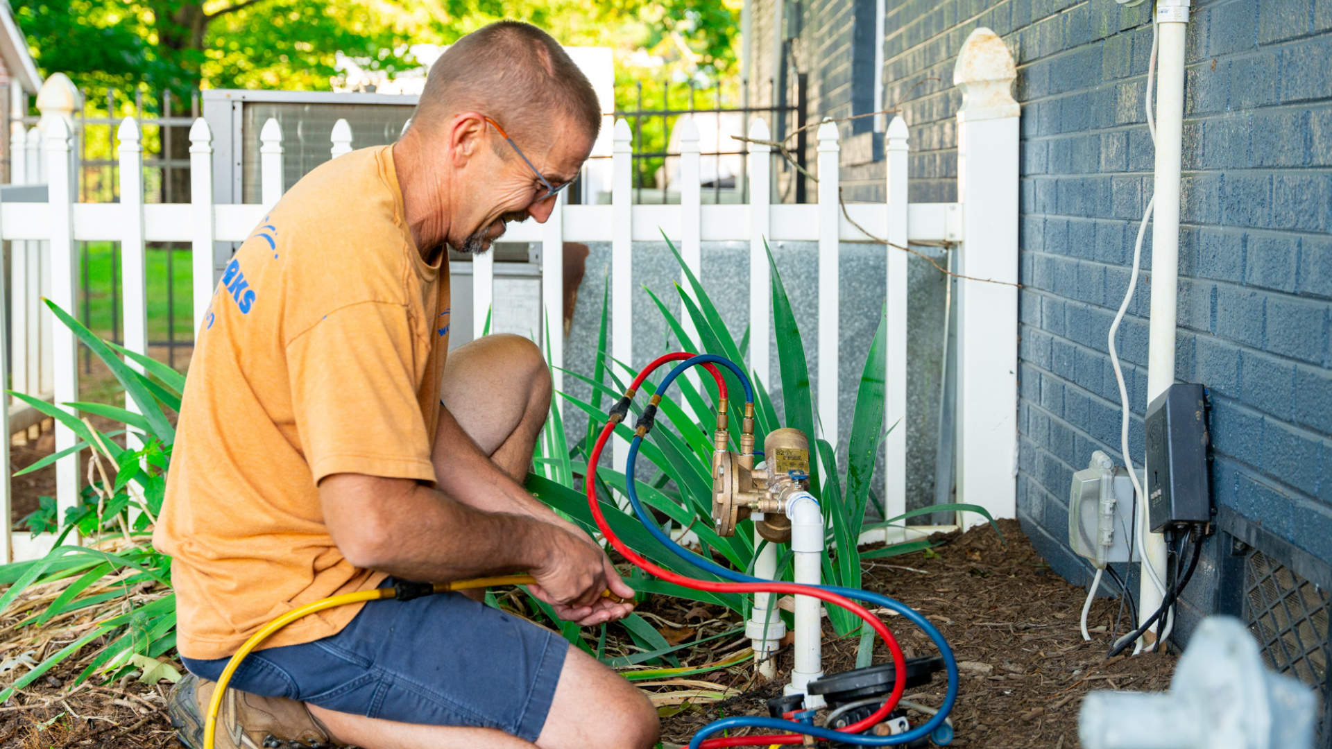 A man working on a garden irrigation system with pipes and hoses near a house with a blue brick wall, a white fence, and green plants.