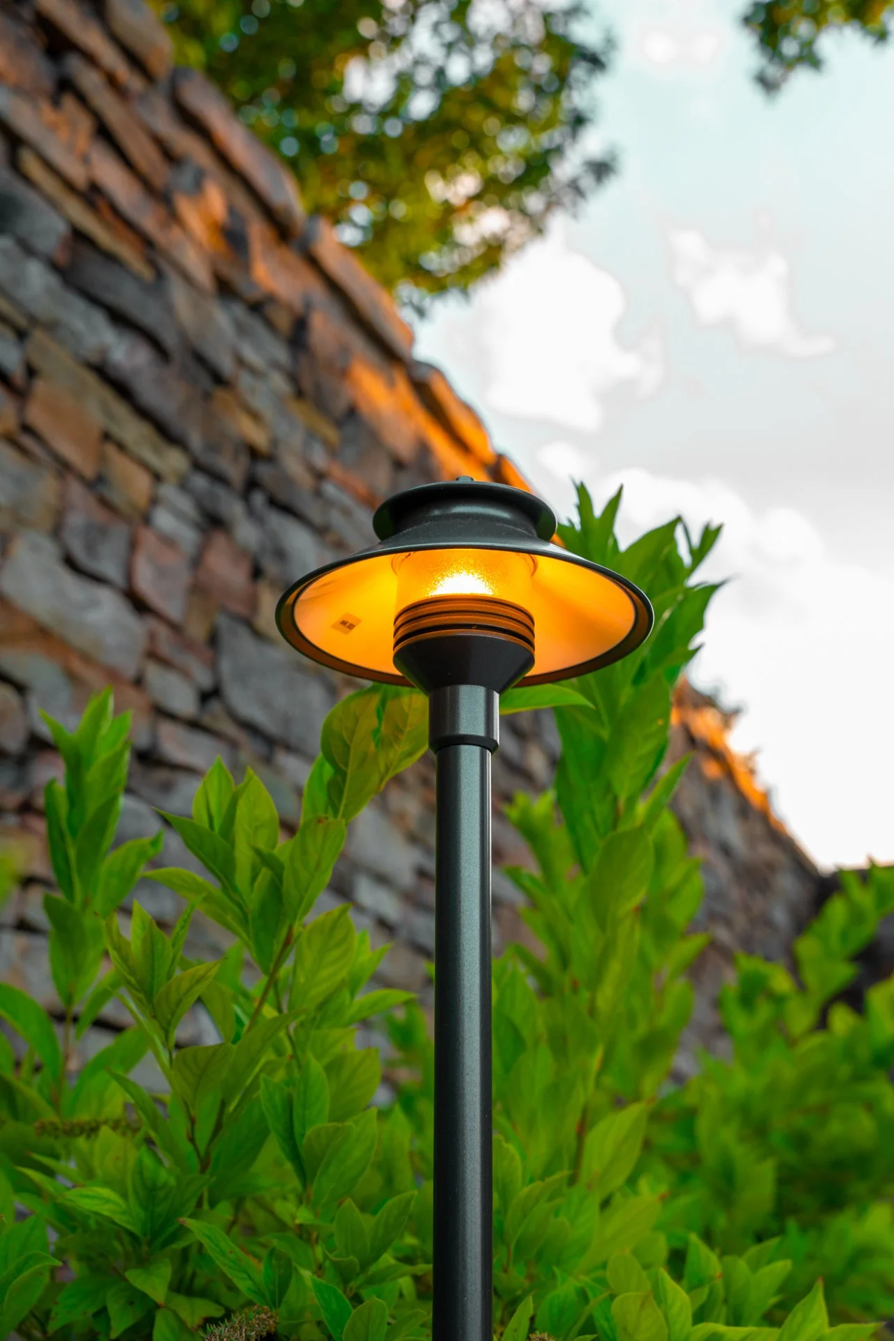 Outdoor garden lamp with a yellow-orange glow, surrounded by green foliage, against a brick wall and a partly cloudy sky.