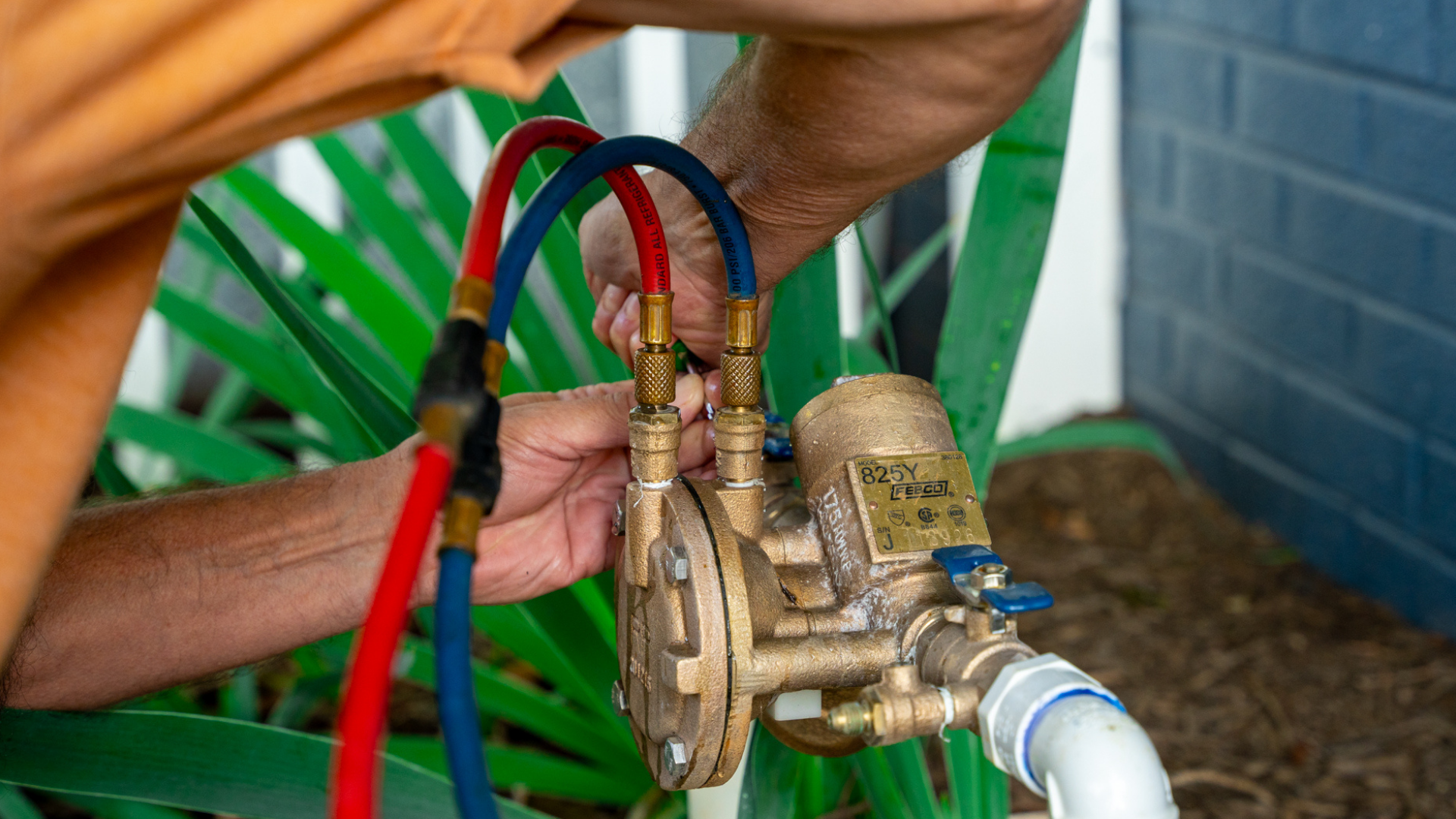Close-up of a person's hands working on a brass valve connected to red and blue hoses, with greenery and a blue wall in the background.