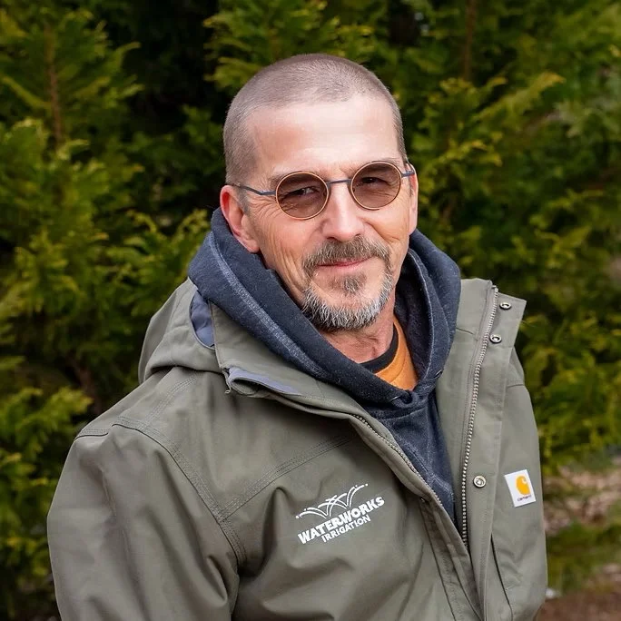 A middle-aged man with glasses and a trimmed beard, wearing a gray Carhartt jacket and a navy hoodie, standing outdoors with green trees in the background.
