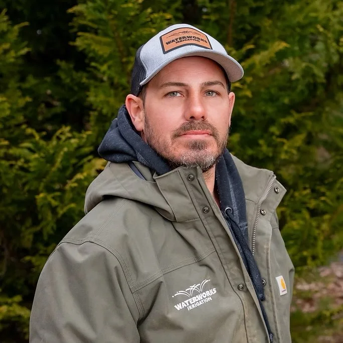 Man wearing a gray trucker hat, gray waterproof jacket, and hoodie outdoors with greenery in the background.
