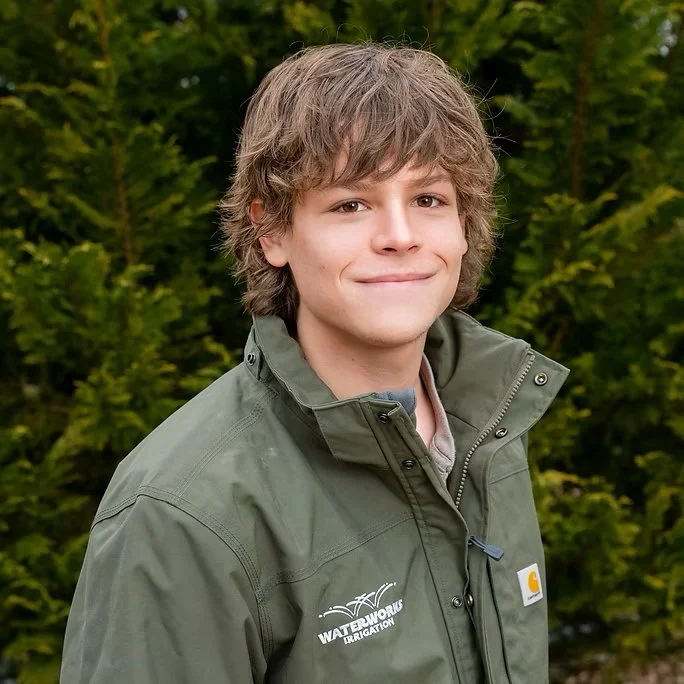 Young man outdoors wearing an olive green Carhartt jacket with WaterWorks Irrigation logo, standing in front of greenery.