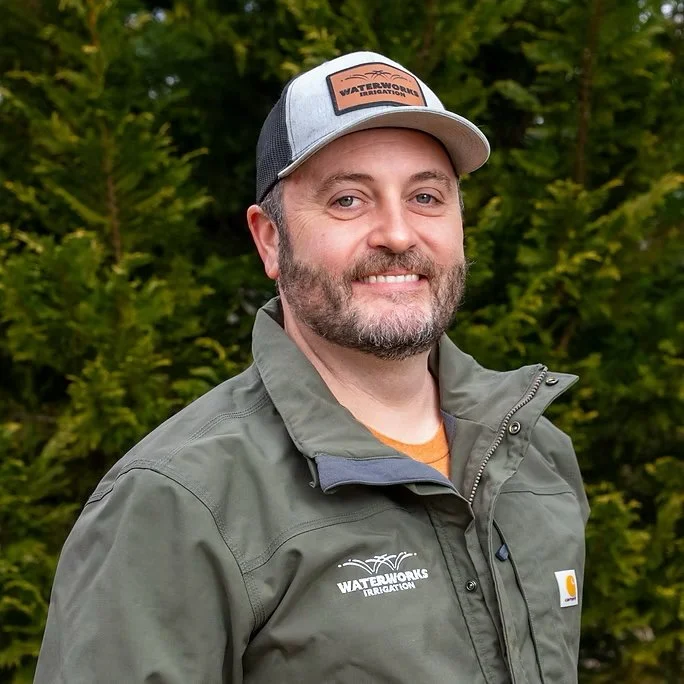 A smiling man outdoors wearing a gray cap with a Waterworks Irrigation logo, a gray jacket with the same logo, and an orange shirt underneath, standing in front of green trees.